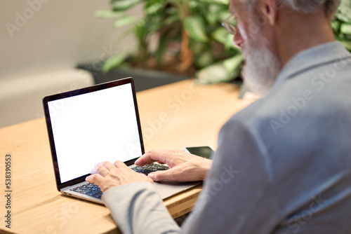 Old senior adult business man teacher typing using laptop with mock up white blank screen template remote learning or working online sitting at office desk. Over shoulder view. Computer mockup