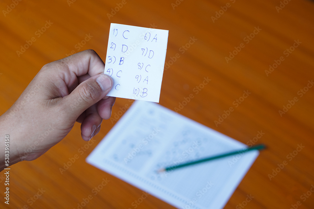 Naklejka premium Closeup student's hand holds small piece of paper with answers during doing the test on pape . Concept : Cheat the test. Dishonest behaviour. Education assessment. 