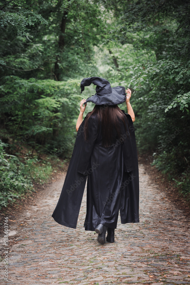 A witch in a black cloak with hat in a dark forest A witch in a black cloak with hat in a dark forest