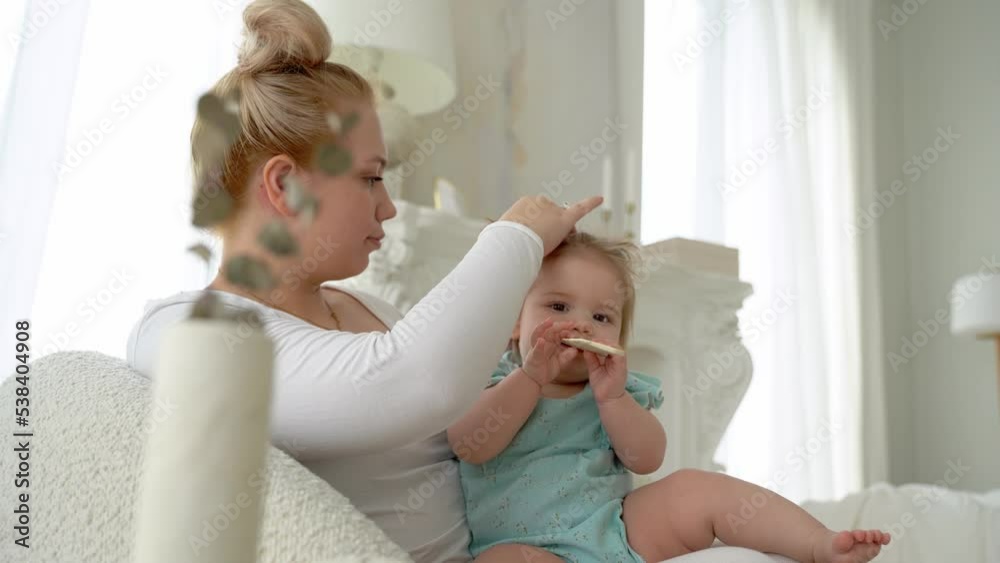 Mother combing hair her baby girl, tie ponytails. Daughter and mummy enjoying time together, smiling. Child care, domestic life routine, healthy lifestyle Mother combing hair her baby girl, tie ponytails. Daughter and mummy enjoying time together, smiling. Child care, domestic life routine, healthy lifestyle