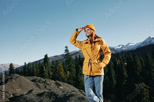 Woman with red hair hiker in yellow raincoat and cap standing on the mountain hands up happiness with a view of snowy mountains travel in winter and hiking in the mountains in the sunset freedom