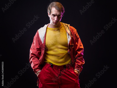 a young muscular man in a bright outfit poses in a pretentious pose, style and grotesque. Attractive guy with pumped-up arms in a mesh T-shirt and red pants, photo in smoke photo