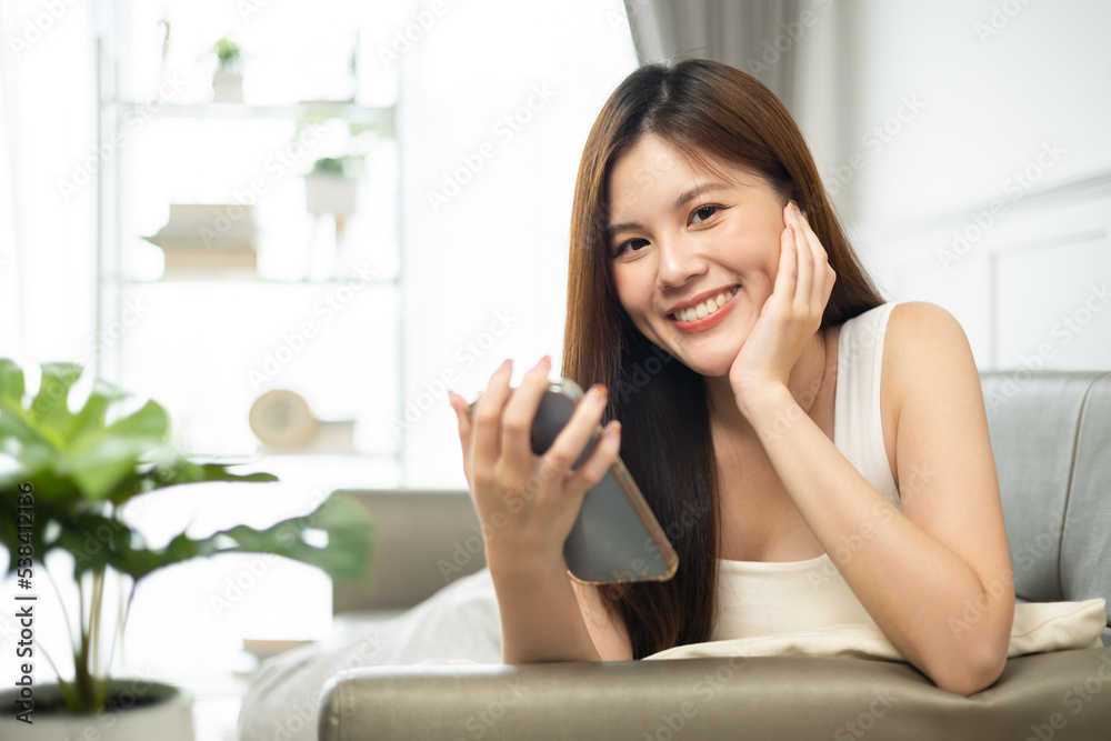 Happy and cheerful woman is using a phone sitting on a sofa.Asian young woman looking mobile phone celebrating with good news or discount voucher for shopping online at home. Happy and cheerful woman is using a phone sitting on a sofa.Asian young woman looking mobile phone celebrating with good news or discount voucher for shopping online at home.