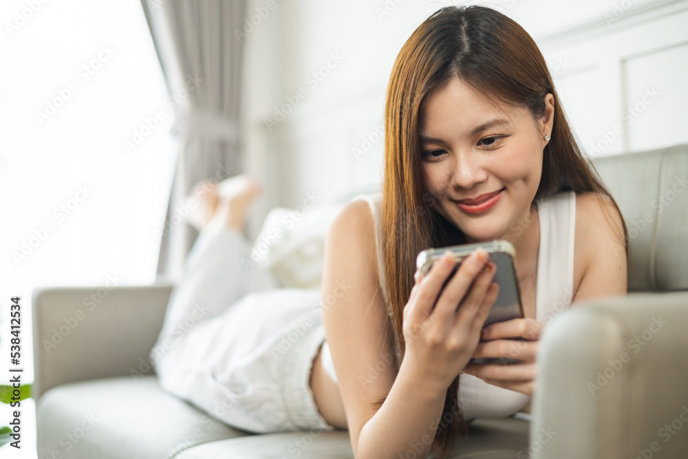 Happy and cheerful woman is using a phone sitting on a sofa.Asian young woman looking mobile phone celebrating with good news or discount voucher for shopping online at home. Happy and cheerful woman is using a phone sitting on a sofa.Asian young woman looking mobile phone celebrating with good news or discount voucher for shopping online at home.