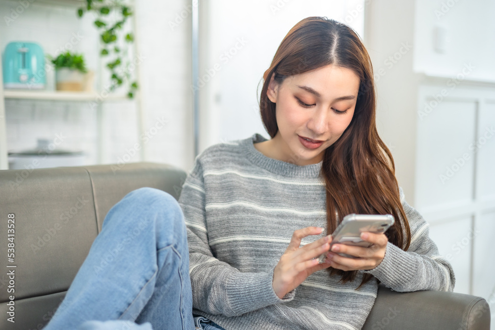 Happy and cheerful woman is using a phone sitting on a sofa.Asian young woman looking mobile phone for shopping online at home. Happy and cheerful woman is using a phone sitting on a sofa.Asian young woman looking mobile phone for shopping online at home.