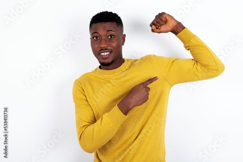 Tablou pe pânză Smiling young handsome man wearing yellow sweater over white background raises hand to show muscles, feels confident in victory, strong and independent