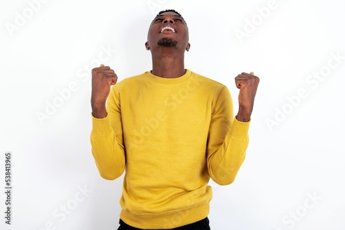 young handsome man wearing yellow sweater over white background looks with excitement up, keeps hands raised, notices something unexpected.