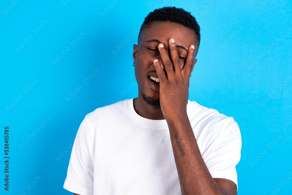 young handsome man wearing white T-shirt over blue background makes face palm and smiles broadly, giggles positively hears funny joke poses young handsome man wearing white T-shirt over blue background makes face palm and smiles broadly, giggles positively hears funny joke poses