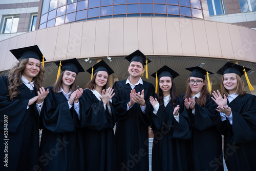 Seven graduates in robes stand in a row and clap outdoors.