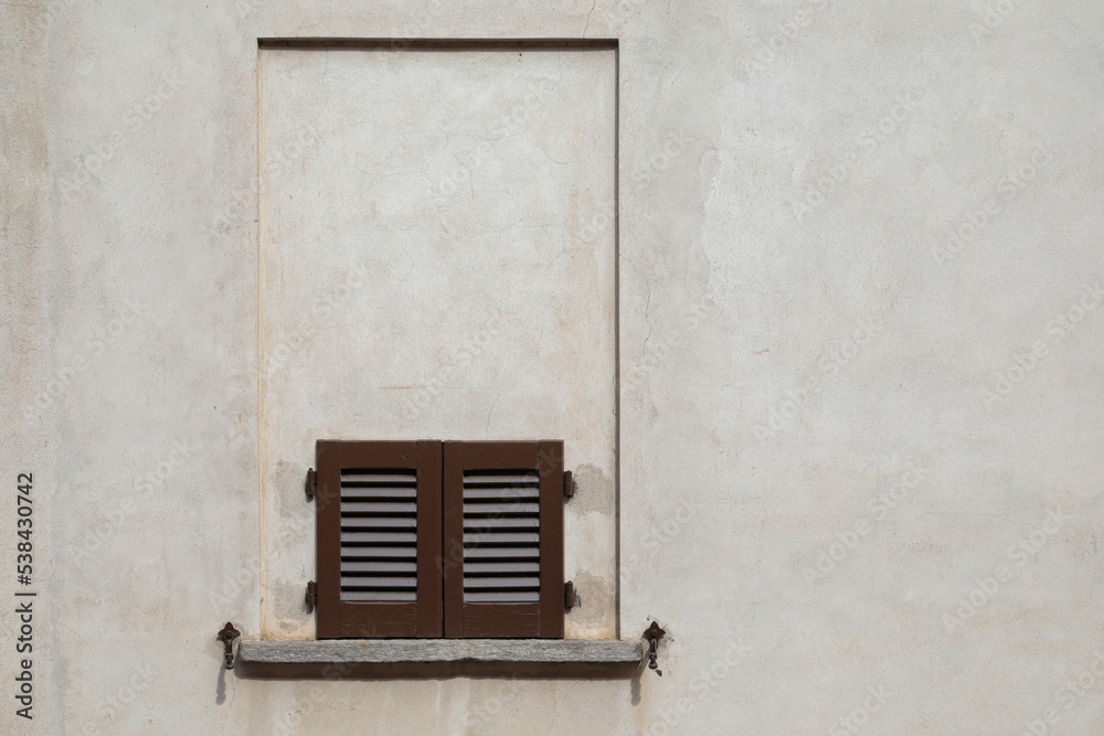 Fototapeta premium Bright stone wall of a house with a bricked up window, and a small window with brown wooden shutter