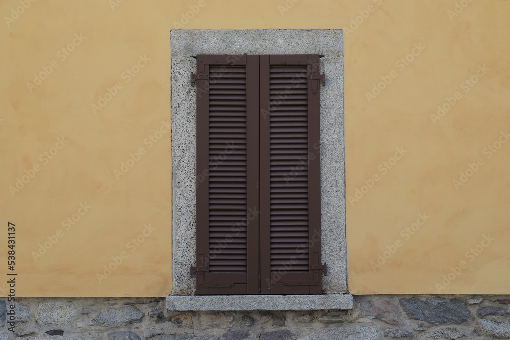 Window with brown wooden shutters on a yellow cement wall and grey stone brick wall, grey frame around the window with windowsill, 