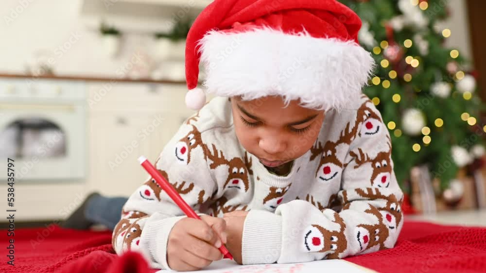 child boy in a santa hat lying on a plaid smiles dreamily and writes a letter to Santa on a white sheet - a wish list on the background of a Christmas tree. child boy in a santa hat lying on a plaid smiles dreamily and writes a letter to Santa on a white sheet - a wish list on the background of a Christmas tree.