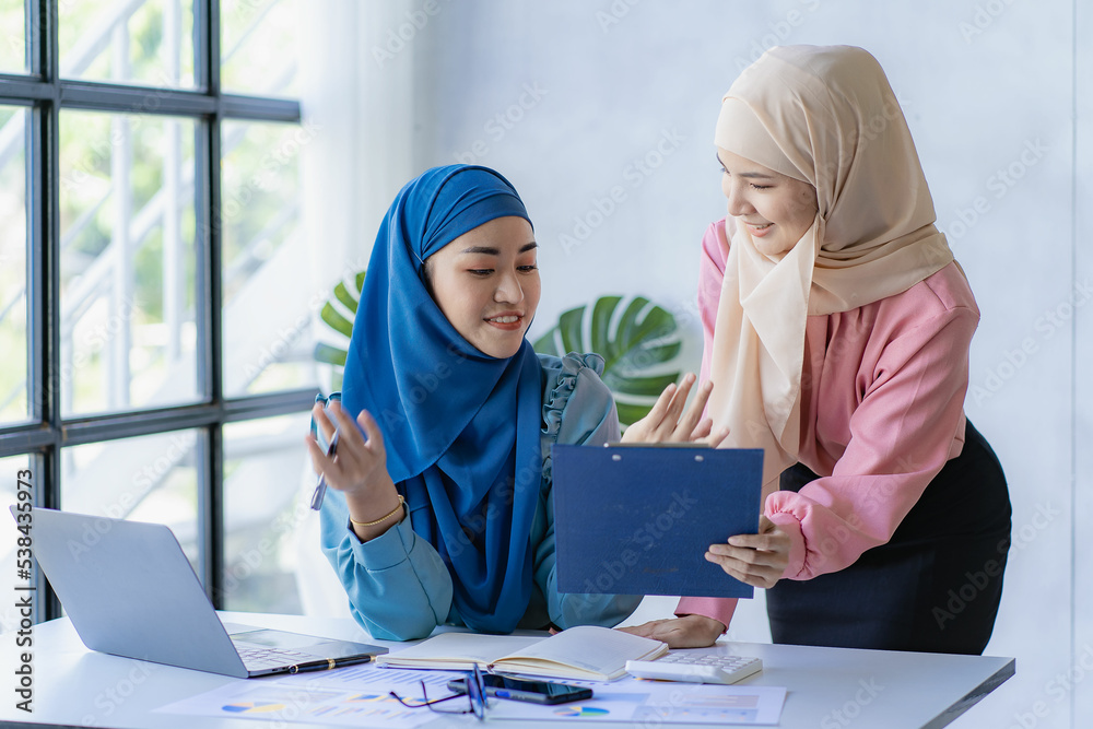 Two beautiful Asian Muslim women wearing headscarves working in the office with laptops with financial graphs.
Muslim businesswoman in traditional dress working and talking in office meeting