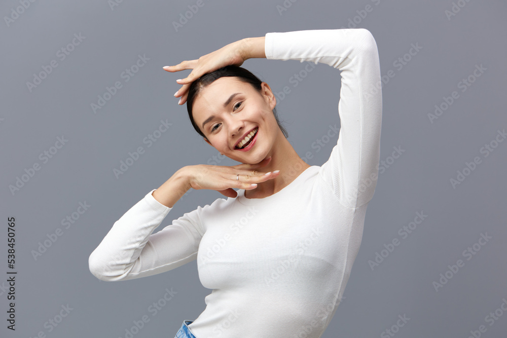 an excited woman stands on a gray background in a white T-shirt, raised one hand above her head, and lowered the other under her chin, gently looking away