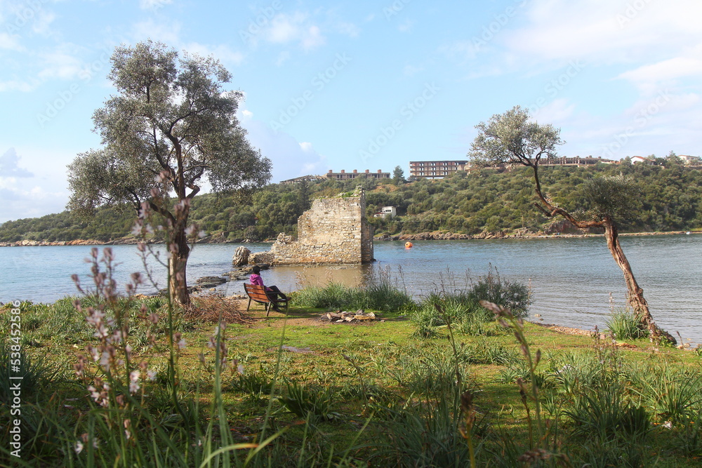 The woman watches the view of the ancient castle alone on the bench. Ruined and restored castle at Iasos Turkey on the Aegean Sea near Bodrum The woman watches the view of the ancient castle alone on the bench. Ruined and restored castle at Iasos Turkey on the Aegean Sea near Bodrum