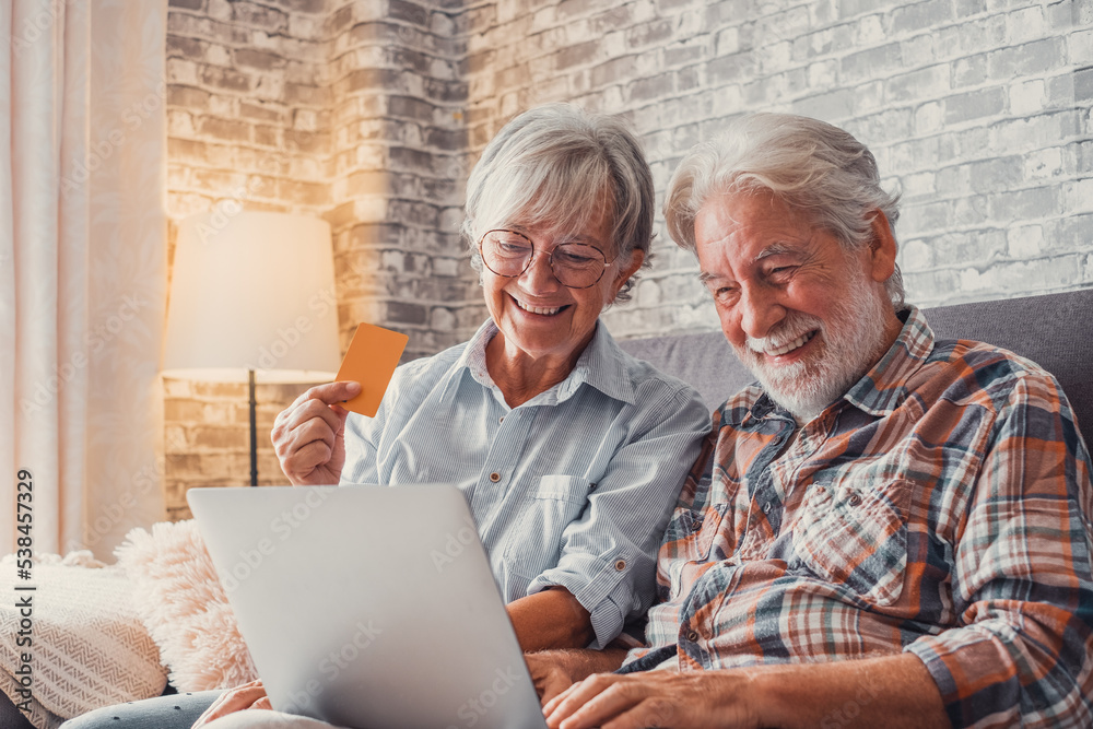 Portrait of couple of cute two happy and excited seniors buying and spending money shopping online with laptop on the sofa at home. Using credit card together buying on internet. Portrait of couple of cute two happy and excited seniors buying and spending money shopping online with laptop on the sofa at home. Using credit card together buying on internet.