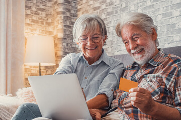Portrait of couple of cute two happy and excited seniors buying and spending money shopping online with laptop on the sofa at home. Using credit card together buying on internet.