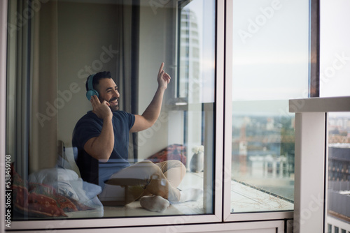  Optimistic man in simple clothes sitting on the balcony at home listening to music and looking away with a smile using wireless headphones