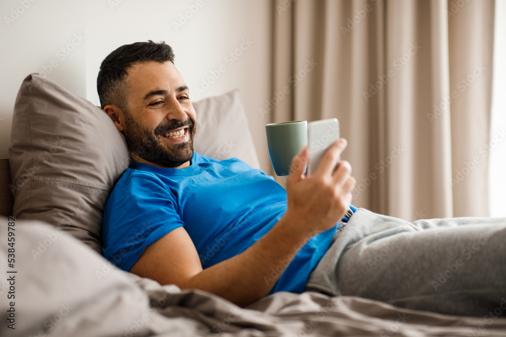 Happy man in blue pajamas lies in bed alone, smiles and reads a text message, close up, side view, copy space Happy man in blue pajamas lies in bed alone, smiles and reads a text message, close up, side view, copy space