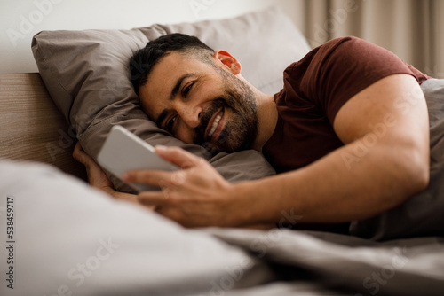 Guy using a smartphone browsing the Internet and sending text messages on the phone lies on the bed at home, free space