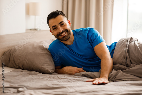 A handsome young man in blue pajamas is lying in bed, touching the empty space next to him and smiling at the camera while waiting for his girlfriend or wife.