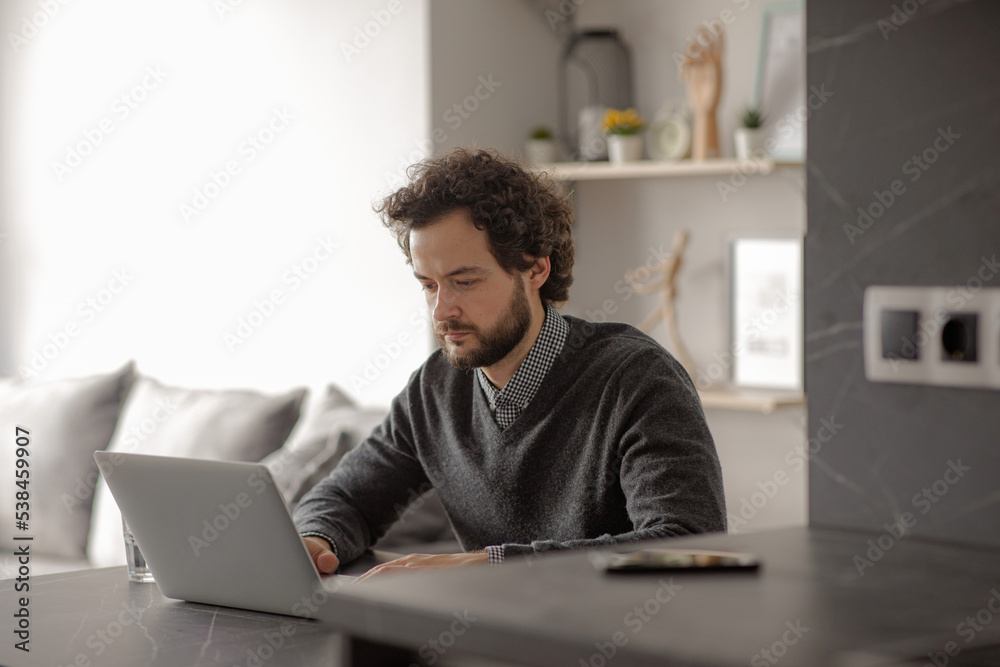 The man works at the leptop. The man is sitting at the table and typing on the computer. The guy works from home in a good mood. Work from home