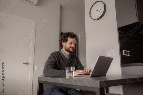 The man works at the leptop. The man is sitting at the table and typing on the computer. The guy works from home in a good mood. Work from home