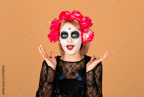 Studio shot of lovely woman wears halloween makeup, dressed in black outfit, red wreath, has zombie image, looks with scaring expression, isolated over beige background, free space for your promotion