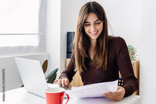 Young smiling pretty adult woman working on laptop while checking home invoices or financial documents
