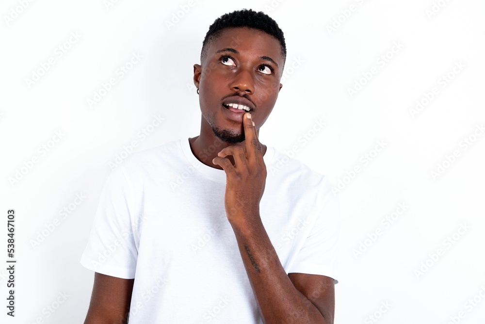 young handsome man wearing white T-shirt over white background with thoughtful expression, looks to the camera, keeps hand near face, bitting a finger thinks about something pleasant. young handsome man wearing white T-shirt over white background with thoughtful expression, looks to the camera, keeps hand near face, bitting a finger thinks about something pleasant.