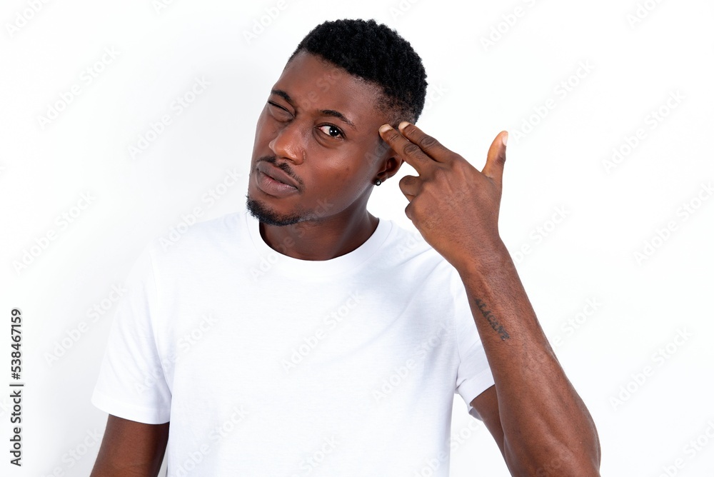 Unhappy young handsome man wearing white T-shirt over white background imitates gun shoot makes suicide gesture keeps two fingers on temples.