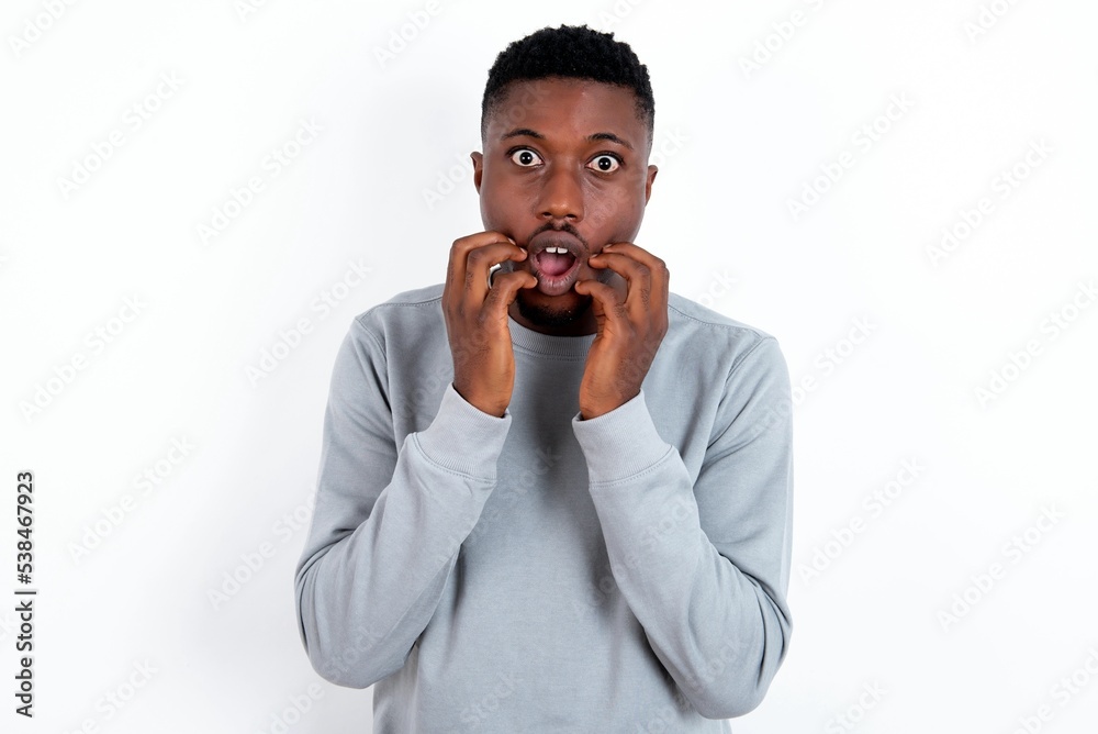 Speechless young handsome man wearing grey sweater over white background keeps hands near opened mouth reacts to shocking news stares wondered at camera Speechless young handsome man wearing grey sweater over white background keeps hands near opened mouth reacts to shocking news stares wondered at camera