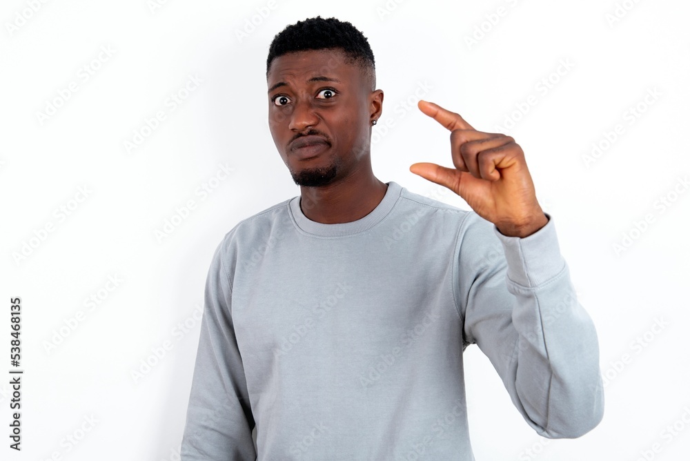 Displeased young handsome man wearing grey sweater over white background shapes little hand sign demonstrates something not very big. Body language concept. Displeased young handsome man wearing grey sweater over white background shapes little hand sign demonstrates something not very big. Body language concept.