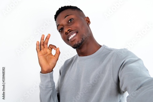 Portrait of pretty cheerful young handsome man wearing grey sweater over white background make selfie show okey symbol
