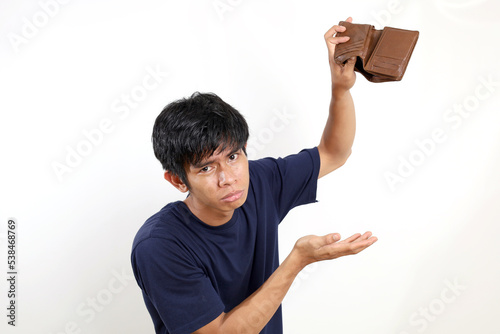 Disappointed young asian man standing while opening his empty wallet. Isolated on white background © SetianingDyah