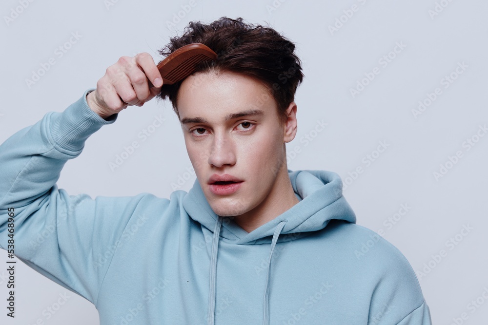 Horizontal portrait photo .a pleasantly smiling young man in a light light blue hoodie, combs his head with a comb place in the background to insert an advertising layout Horizontal portrait photo .a pleasantly smiling young man in a light light blue hoodie, combs his head with a comb place in the background to insert an advertising layout