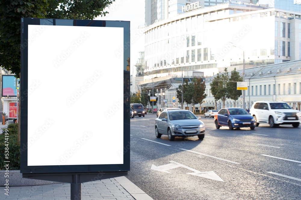 White blank vertical billboard in the city. Bright sun reflects off the office building. Cars drive along the highway. Mock-up.