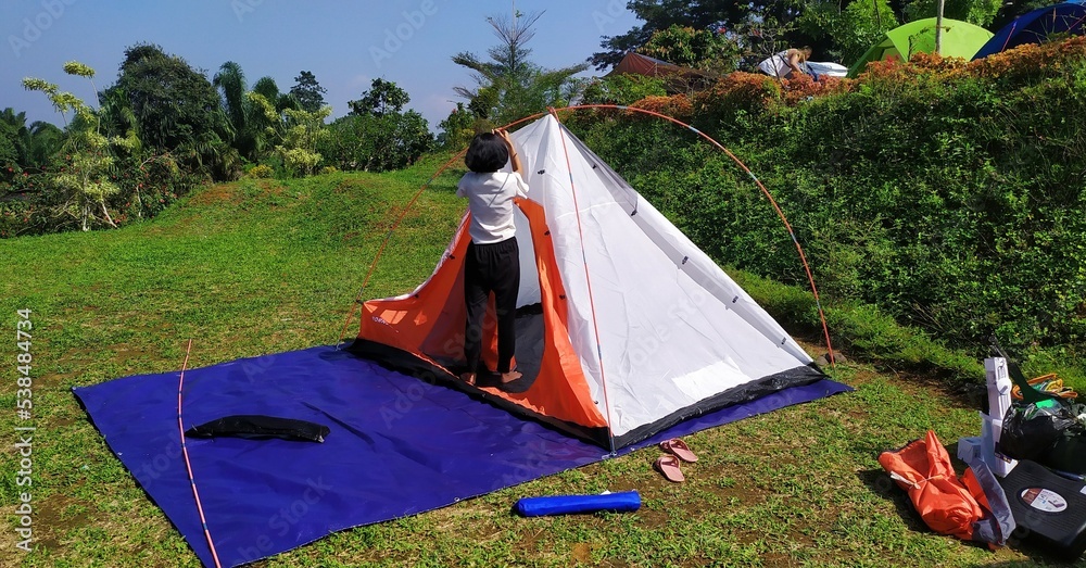 A woman is setting up a white, orange, blue cloth tent on a camping ground during the day in Bogor Indonesia