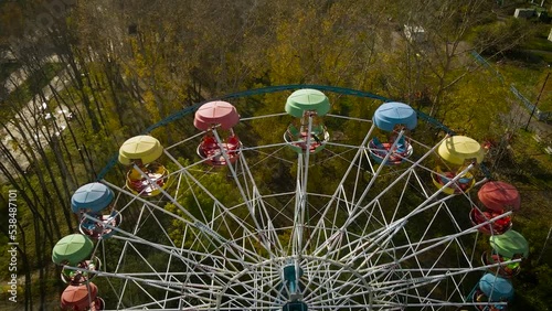 Aerial view of the city ferris wheel with bucket seat in golden autumn amusement park under light blue cloudy sky, drone flying inside
