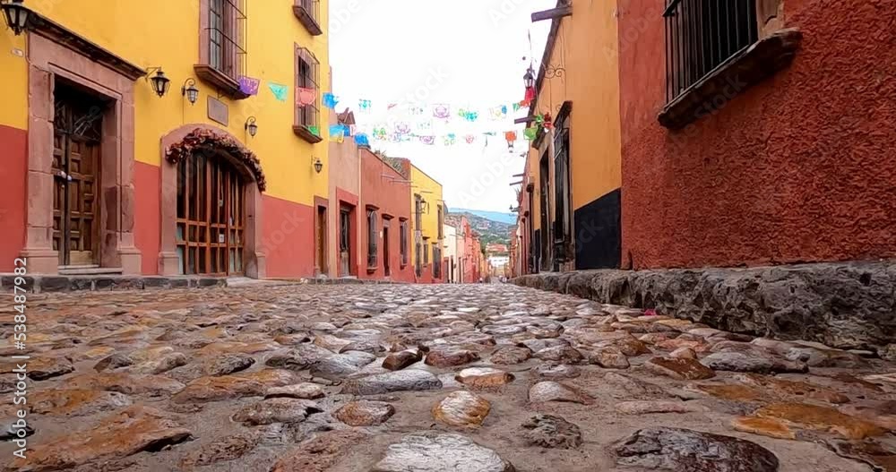 A vibrant cobblestone street in a Mexican town, lined with colorful colonial buildings. Papel picado decorations hang across the street, creating a festive atmosphere under a bright sky.
