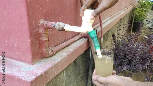 Hand holding and filling glass with less contaminated dirty, rusty and muddy brown drinking water from outdoor wall tap pipe. Indian and African water scarcity, shortage, crisis or impurity concept.