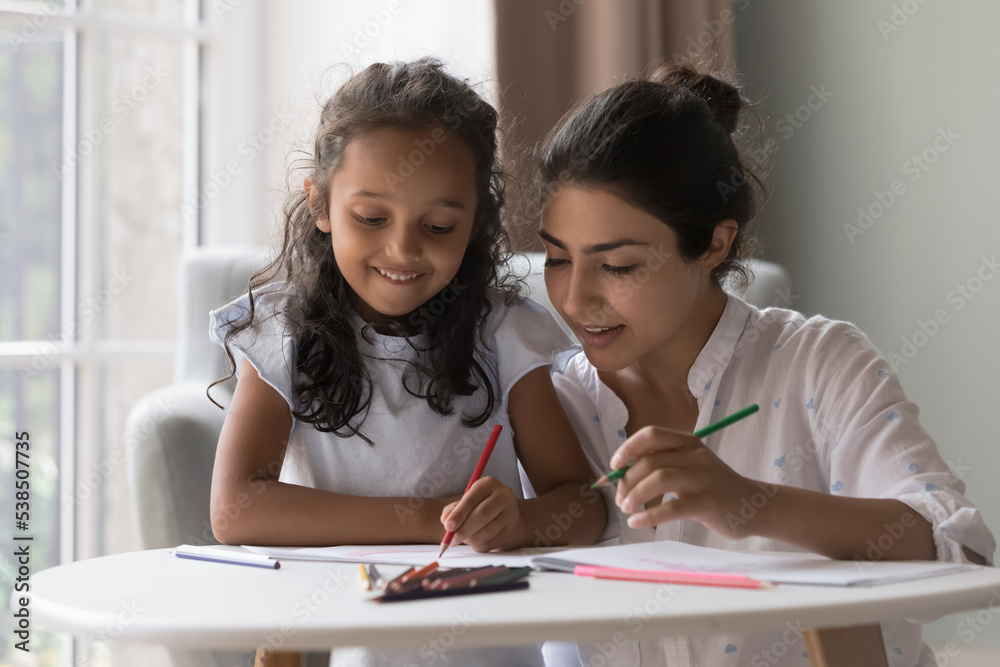 Attractive young Indian mother and little daughter drawing pictures in paper with colored pencils. Loving family enjoy communication and common creative hobby spend leisure at home. Artistic pastime Attractive young Indian mother and little daughter drawing pictures in paper with colored pencils. Loving family enjoy communication and common creative hobby spend leisure at home. Artistic pastime