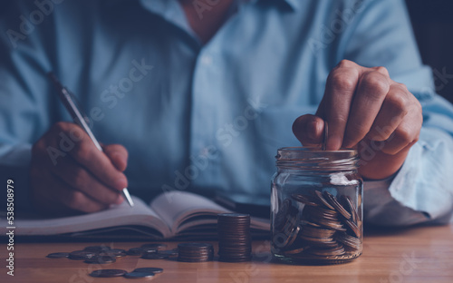 Money saving concept, hand putting coins into the jar and stack coin on wood table. Calculation or count money for saving by record in book for fund, financial investment and loan. 