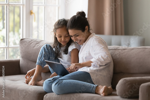 Little cute Indian girl and mom use digital tablet, resting together at home, buying goods through retail services webstore, learn new amusing application seated on cozy sofa, enjoy modern tech usage © fizkes