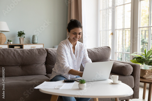 Happy Indian woman making personal budget earning and expenses analysis at home, smile enjoy enough money, financial growth and success, paying bills through e-bank app use laptop. Finances, savings