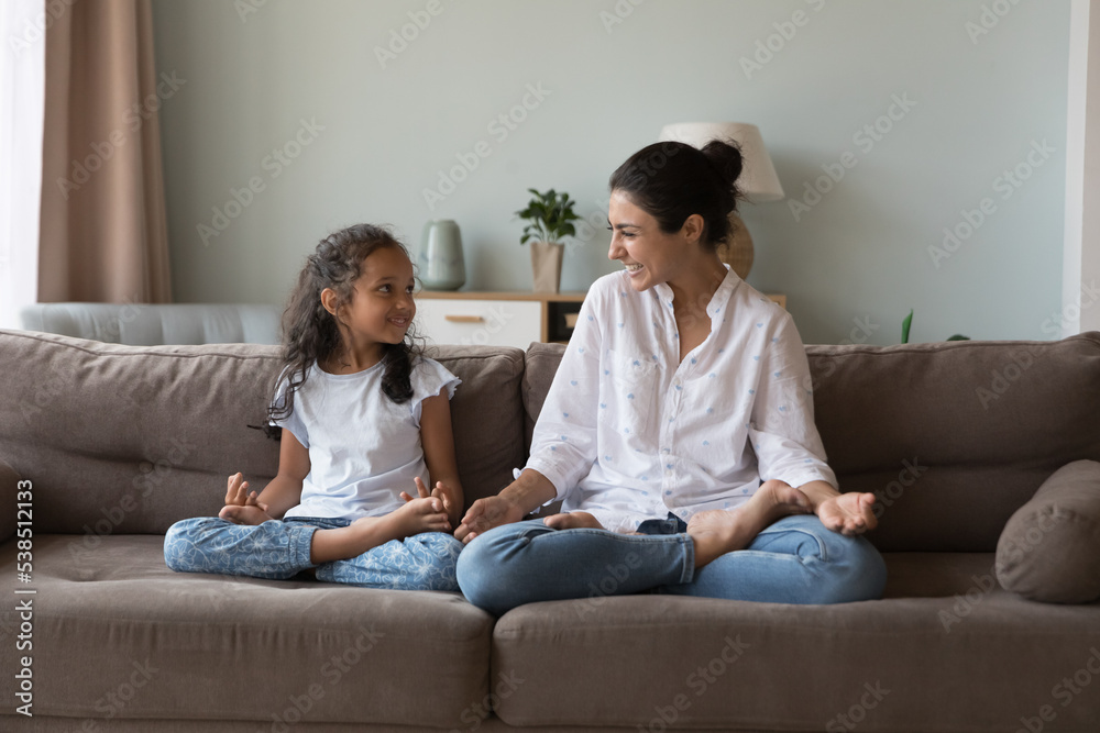 Cheerful Indian woman laughing seated cross-legged on sofa with cute little daughter, loving mother teach her preschooler child good life habit, enjoy yoga training together at modern cozy home. Hobby