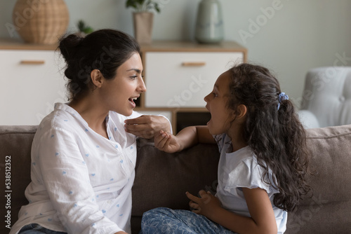 Young Indian mother and cute daughter practicing speaking together, doing vocal exercises seated on sofa at home, therapist logopaedist correct pronunciation of preschooler girl, help to stutter kid photo