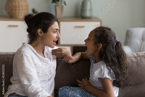 Young Indian mother and cute daughter practicing speaking together, doing vocal exercises seated on sofa at home, therapist logopaedist correct pronunciation of preschooler girl, help to stutter kid