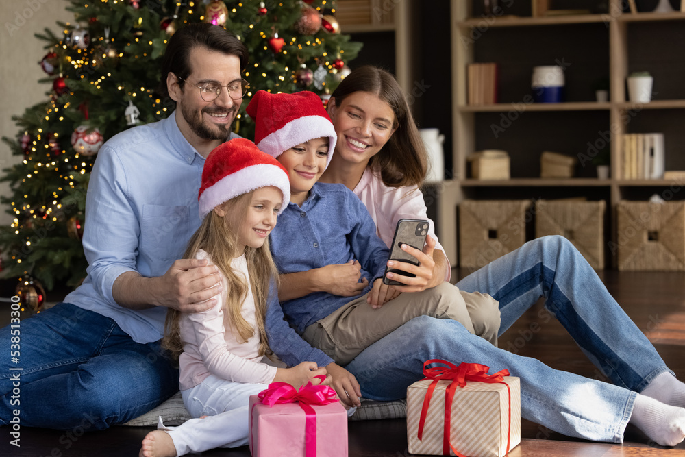 Cheerful two kids in Santa hats and happy parents talking on video phone call, taking family selfie, resting at Christmas tree, gift boxes, enjoying traditional celebration, winter holidays at home