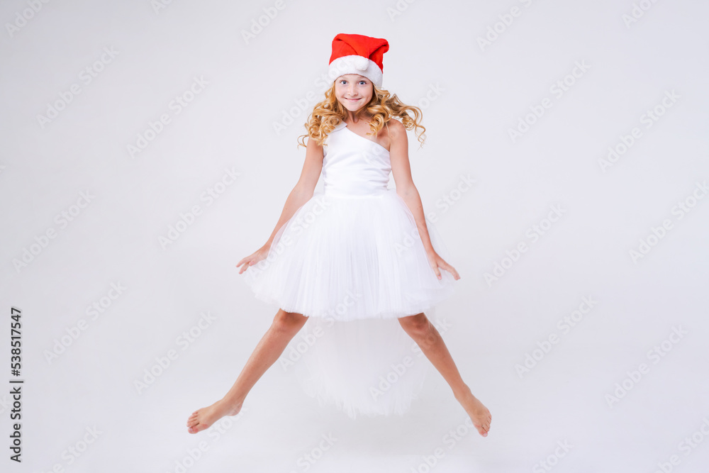 Little joyful girl in red Santa hat and white beautiful dress jumps from happiness and anticipation of the winter holidays of Christmas and New Year on a white background in the studio. Little joyful girl in red Santa hat and white beautiful dress jumps from happiness and anticipation of the winter holidays of Christmas and New Year on a white background in the studio.